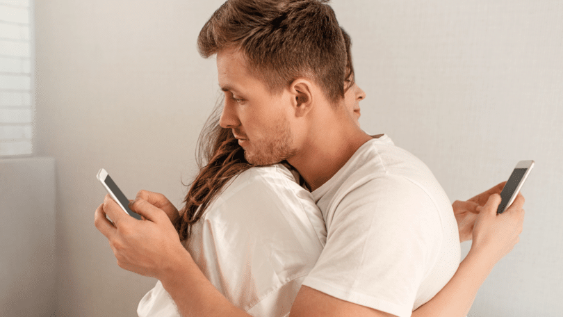 Casal abraçados na cama, enquanto usam os celulares. Conceito de traição.