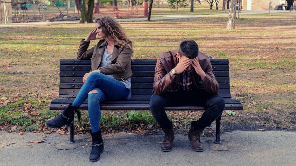 Um casal sentado em um banco de uma praça. Ambos parecem estar intrigados.