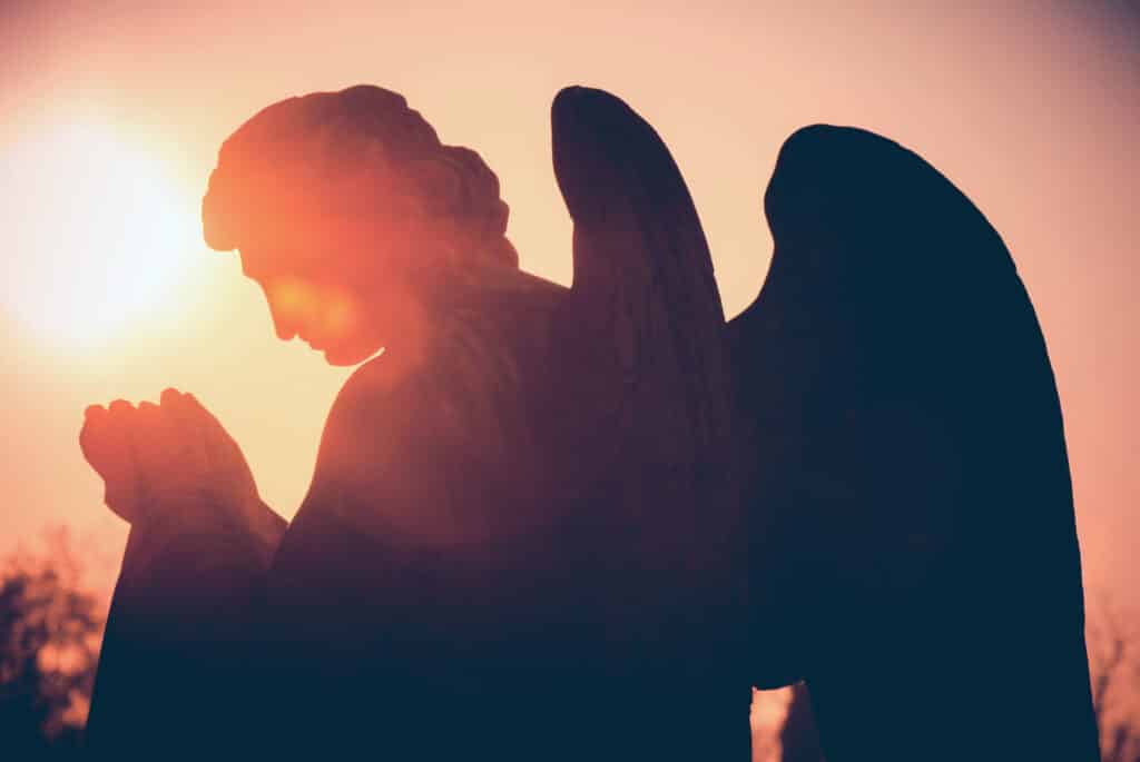 Silhueta da estátua de um anjo com as mãos juntas em oração. Ao fundo, o Sol brilha no céu.