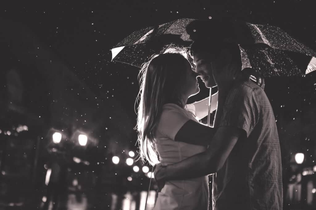 Homem e mulher quase se beijando sob um guarda chuva. É noite e algumas gostas de chuva caem ao seu redor.