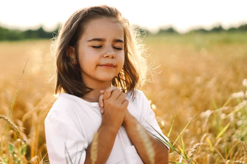 Garotinha orando com as mãos unidas e os olhos fechados. Ela está ao ar livre, em meio a uma plantação, e seu semblante é sereno.