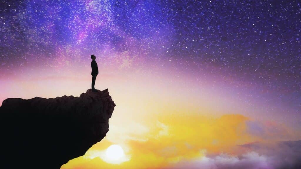 Uma pessoa, à beira de um precipício, a contemplar um céu estrelado e auroreado.
