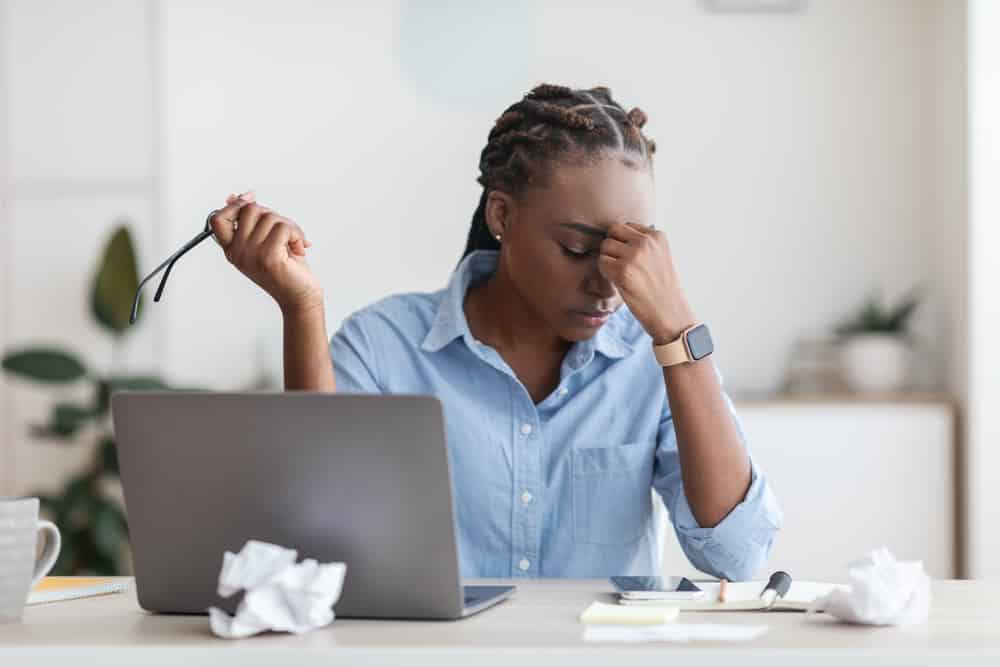 Mulher negra em escritório com os olhos fechados e mão na cabeça, em expressão de estresse