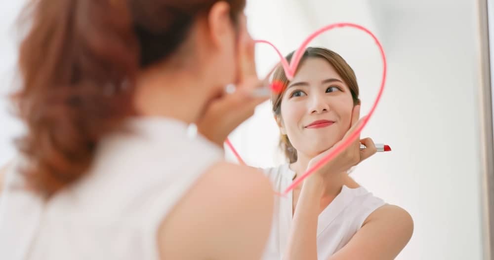 Mulher com um batom vermelho na mão se olhando feliz para um espelho com um coração desenhado em seu reflexo.