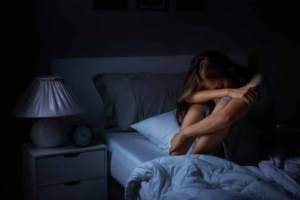 Mulher sentada na cama, em posição fetal, com as luzes do quarto apagadas.