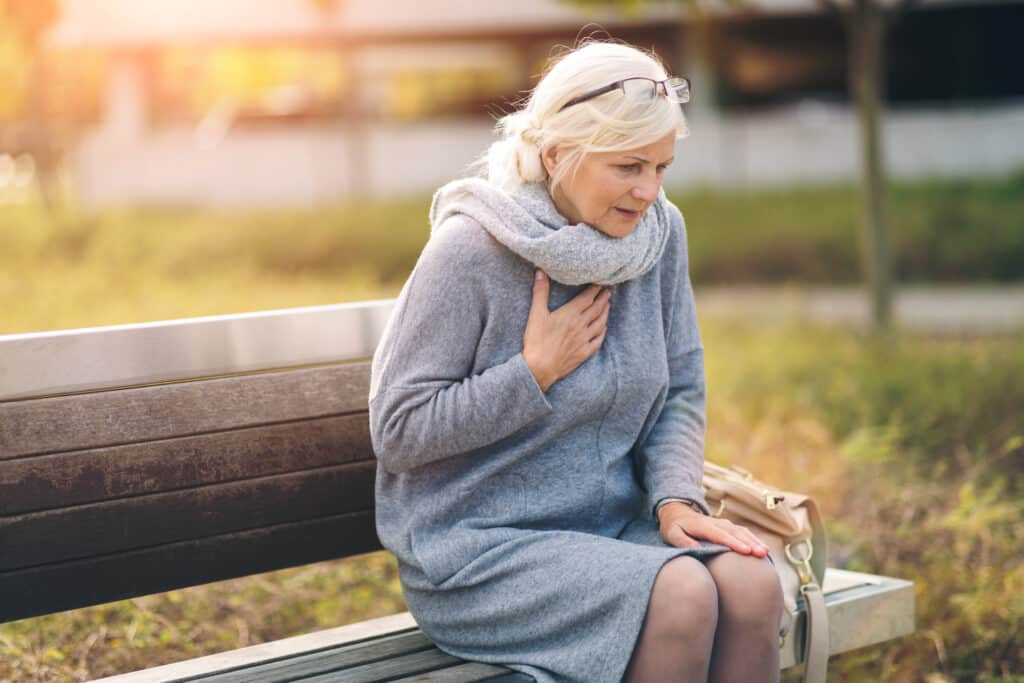 Mulher idosa sentada em um banco de praça, com a mão no coração.