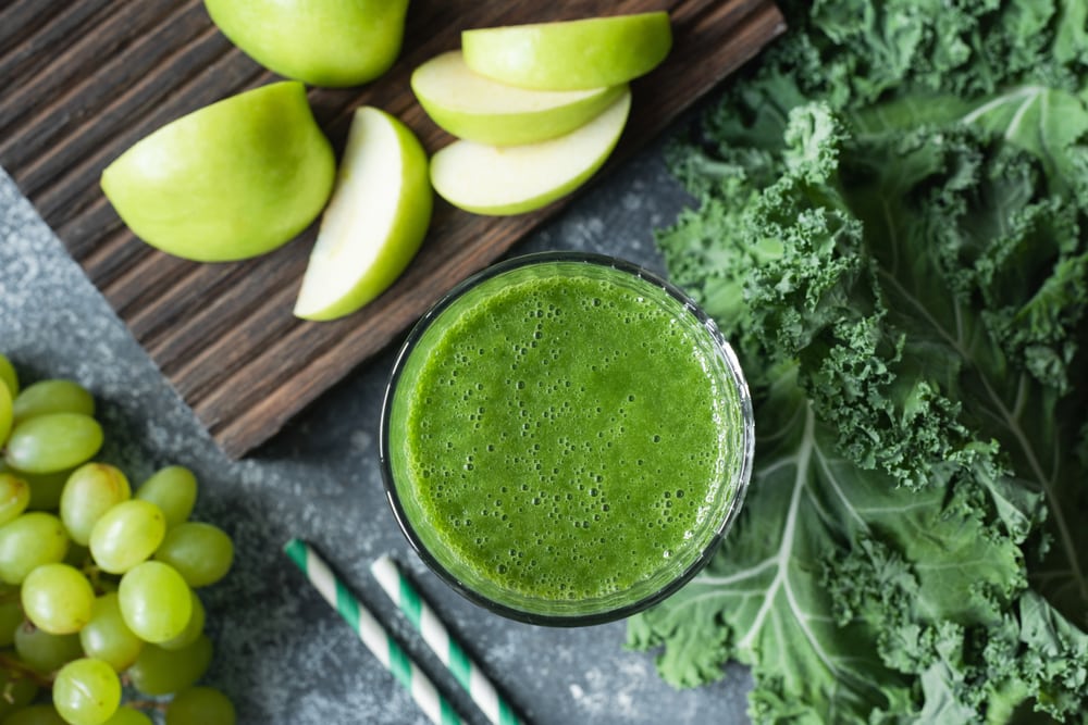 Copo com suco de couve visto de cima. Ao seu lado, folhas de couve, fatias de maçã verde, um cacho de uvas verdes e dois canudos.