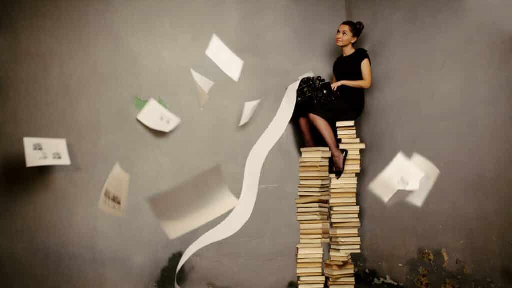 Uma mulher sentada numa pilha de livros. Ela segura, também, uma grande folha de papel.