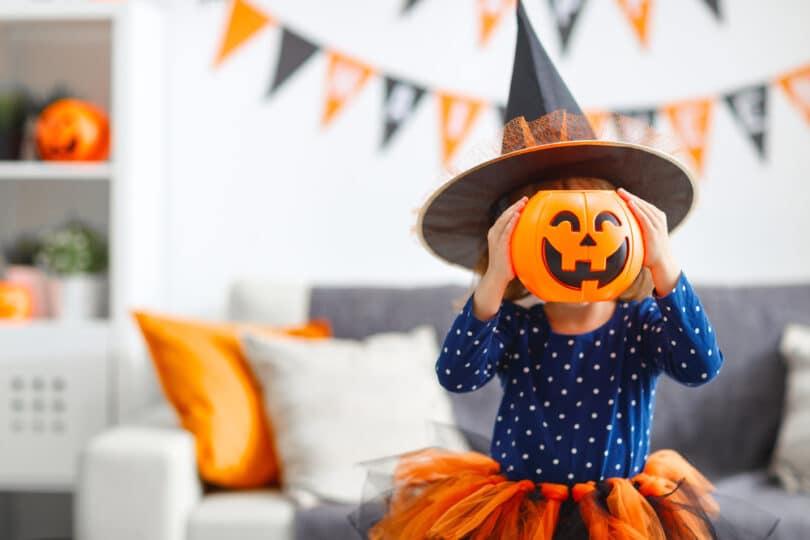 Uma garota fantasiada de bruxa. Ela segura uma abóbora de halloween à altura da cabeça.