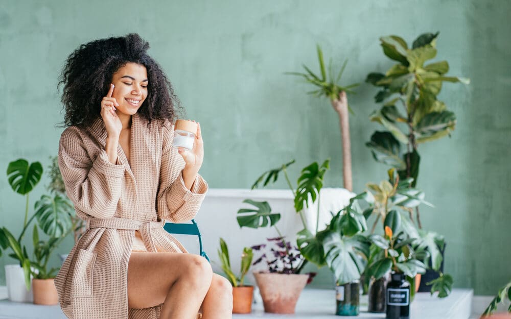 Uma mulher utilizando cosméticos. Ao lado e atrás dela, várias plantas diferentes.