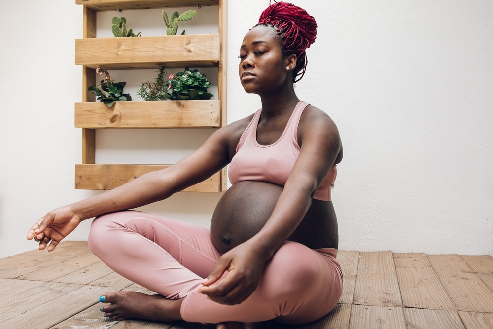 Mulher negra grávida sentada no chão, sobre as pernas cruzadas, em posição de meditação e com os olhos fechados. Ao fundo, apenas algumas plantas sobre prateleiras de madeira.