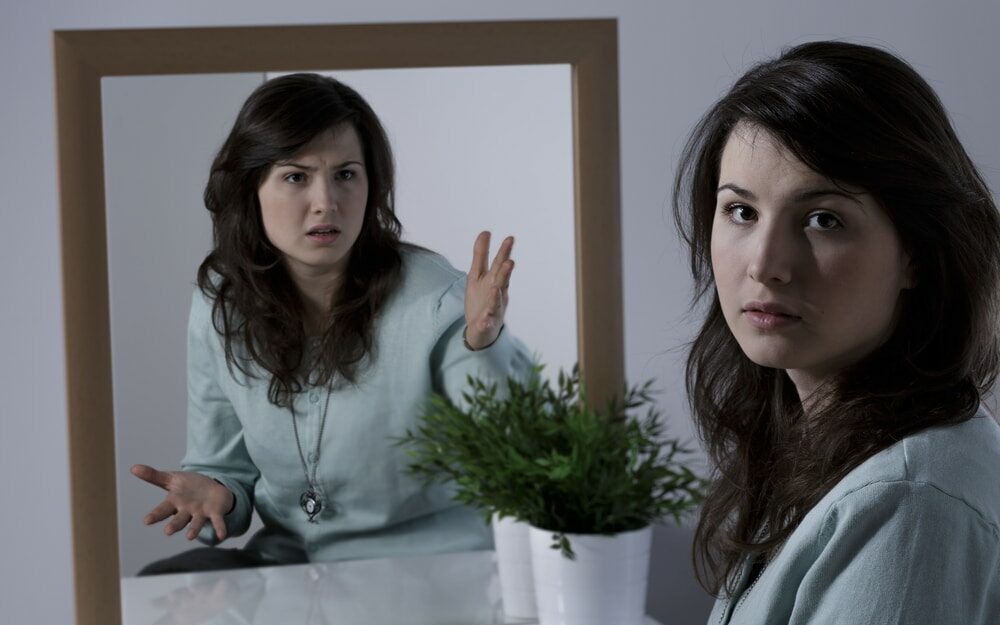 Uma mulher olhando lateralmente. Ao lado, um espelho  a reflete exibindo um semblante raivoso.