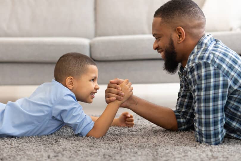 Um homem fazendo uma queda de braço com um garoto. Ambos sorriem.