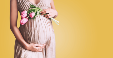 Mulher de vestido longo grávida segurando flores sobre a barriga, em fundo amarelo.