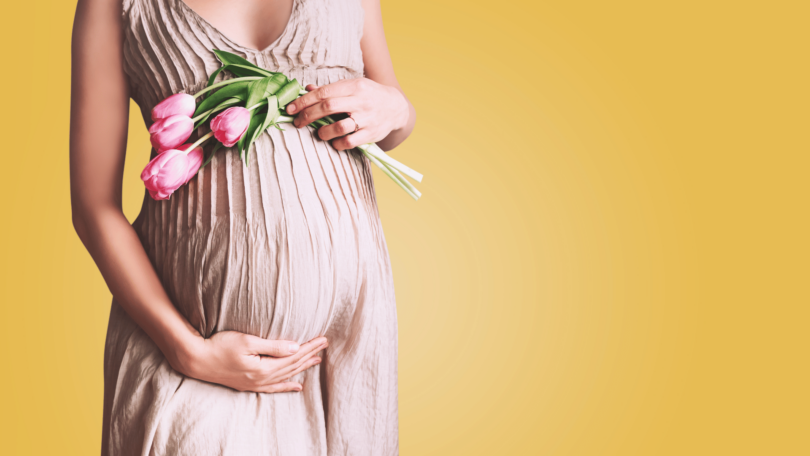 Mulher de vestido longo grávida segurando flores sobre a barriga, em fundo amarelo.
