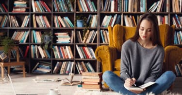 Uma mulher sentada próxima a uma poltrona. Atrás dela, uma grande biblioteca de livros.