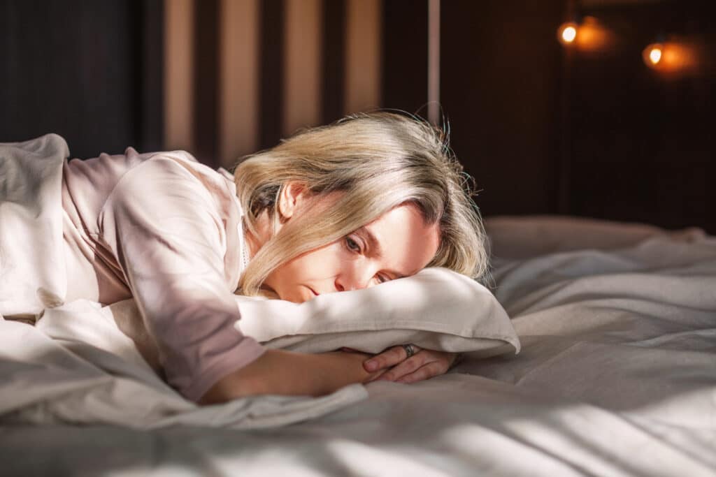 Mulher loira deitada na cama com semblante triste.