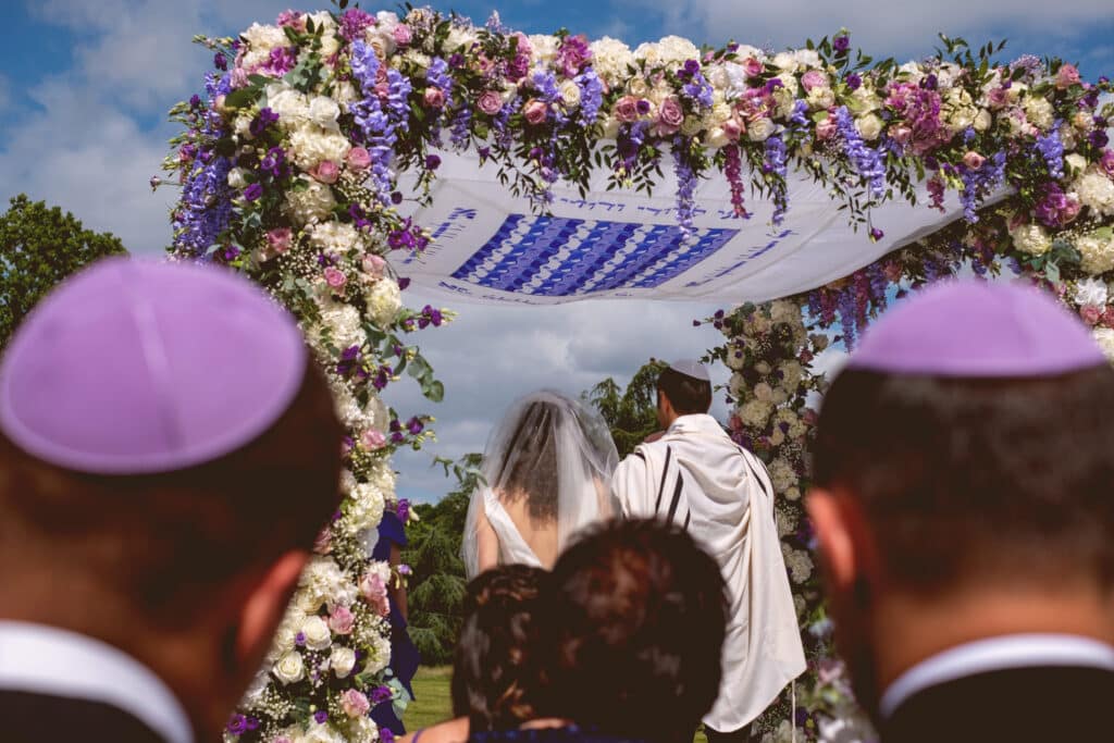 Noivos sob a chupá florida durante a cerimônia de casamento. Seus convidados, ao redor, acompanham o momento.