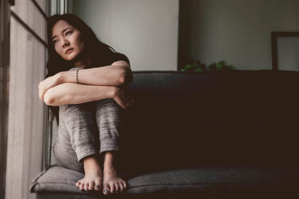 Mulher asiática com semblante triste. Ela está sentada no canto do sofá, abraçando os próprios joelhos, com a cabeça encostada na janela, olhando para fora.