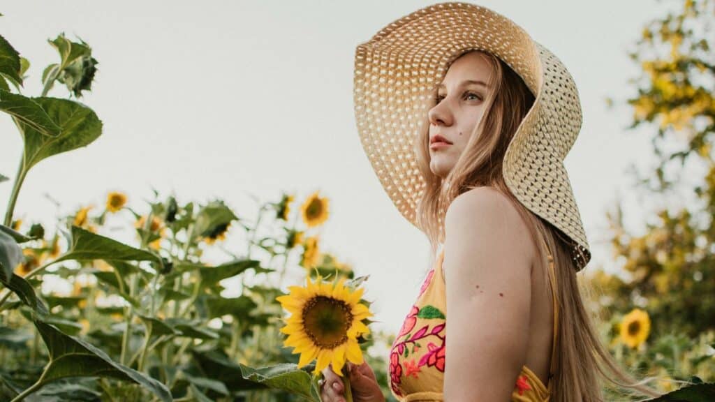 Uma mulher ao meio de um campo florido. Ela segura um girassol. 