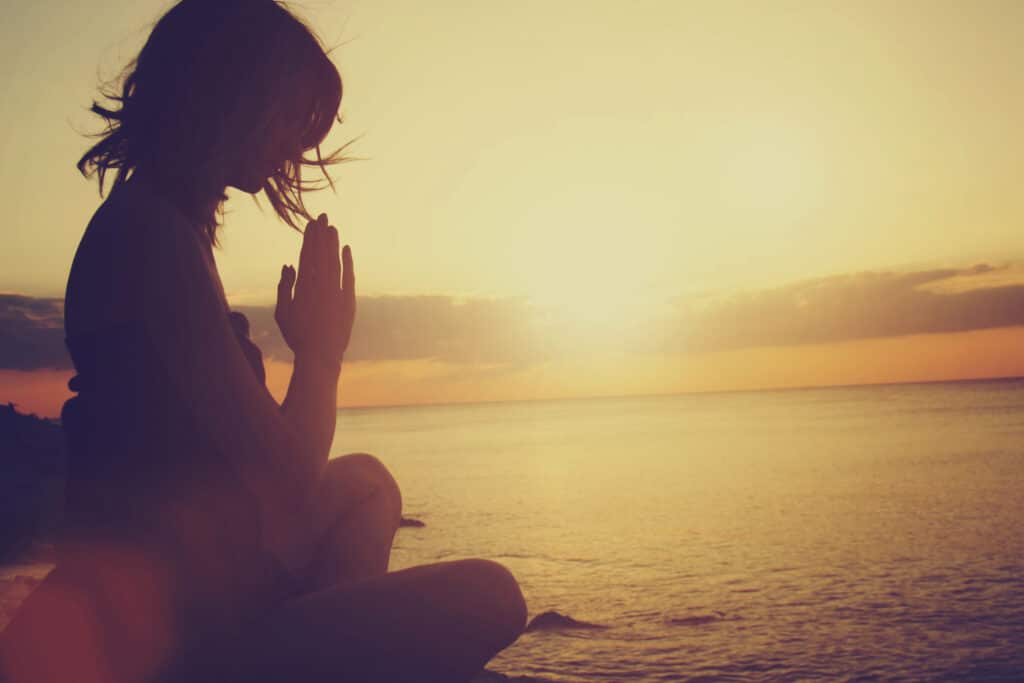 Jovem mulher meditando sentada à beira do mar. Ao fundo, o Sol, entre nuvens, ilumina a paisagem.