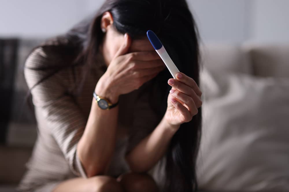 Mulher segurando um teste de gravidez e escondendo o rosto com a outra mão.