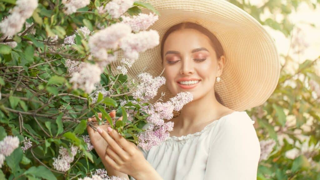 Uma mulher de chapéu sorrindo. Ela segura flores.