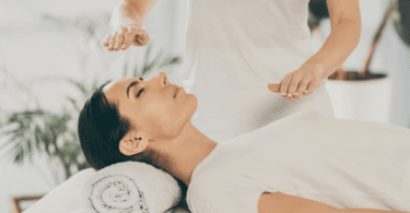 Mulher deitada em uma sessão de Reiki. A terapeuta está com as mãos espalmadas logo acima de sua testa e de seu peito.