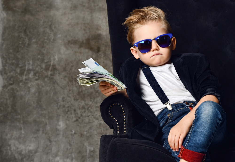 Criança sentada em sofá de couro com óculos escuro fantasiado de magnata com dinheiro na mão 