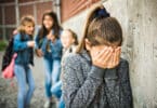 Trio de garotas rindo e apontando para uma menina sozinha encostada na parede com as mãos no rosto chorando