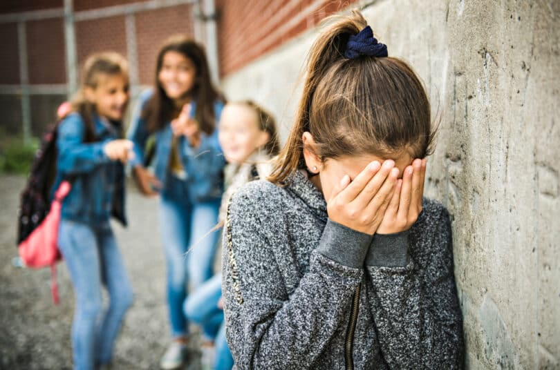 Trio de garotas rindo e apontando para uma menina sozinha encostada na parede com as mãos no rosto chorando