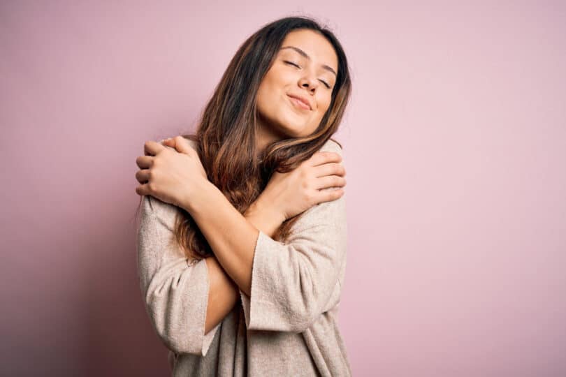 Mulher abraçando a si mesma enquanto sorri de olhos fechados num plano de fundo lilás liso