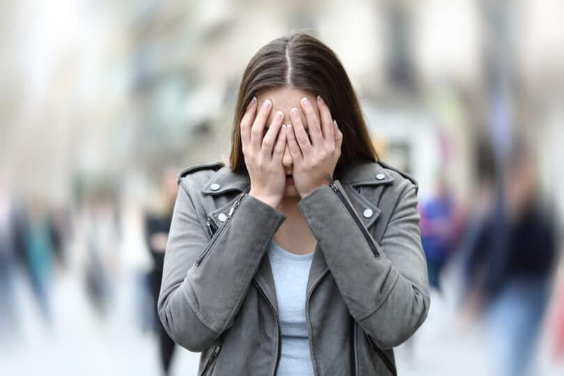Mulher de pé na rua com mãos no rosto e o fundo desfocado indicando tontura ou pavor