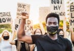 Homem negro parado fazendo o símbolo da resistência com a mão enquanto há pessoas atrás com cartazes em prol do Black Lives Matter