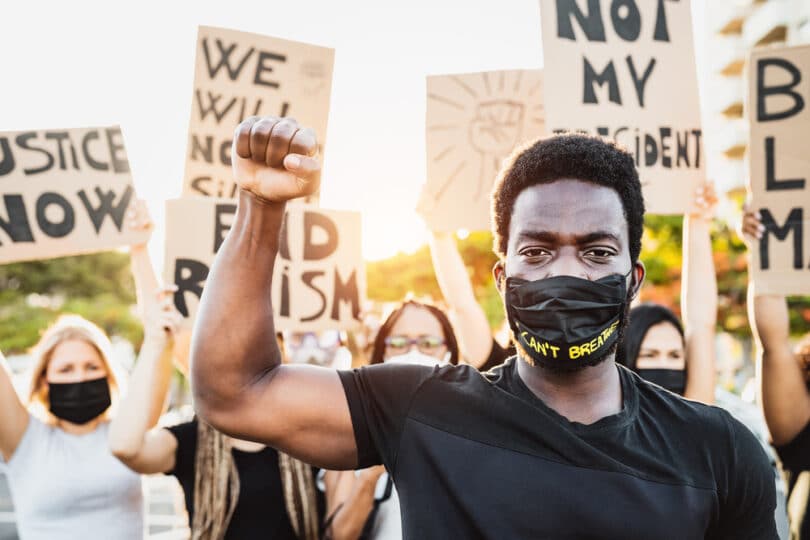 Homem negro parado fazendo o símbolo da resistência com a mão enquanto há pessoas atrás com cartazes em prol do Black Lives Matter