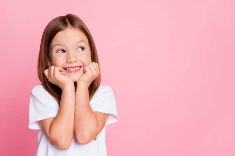 Menina sorrindo com mãos no queixo em fundo liso rosa