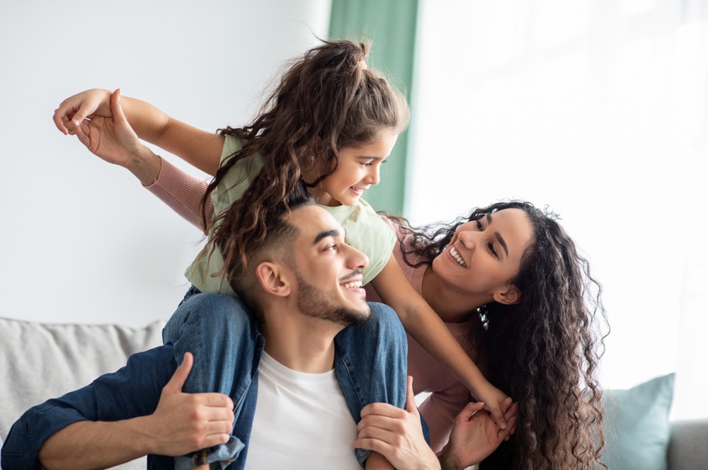 Família feliz unida, criança no ombro do pai de braços abertos e olhando sorridente para mãe que também sorri 