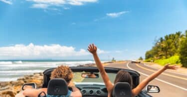 Casal de férias dentro de um conversível aberto na estrada. Na imagem há uma praia no fundo e a mulher está de braços abertos para cima