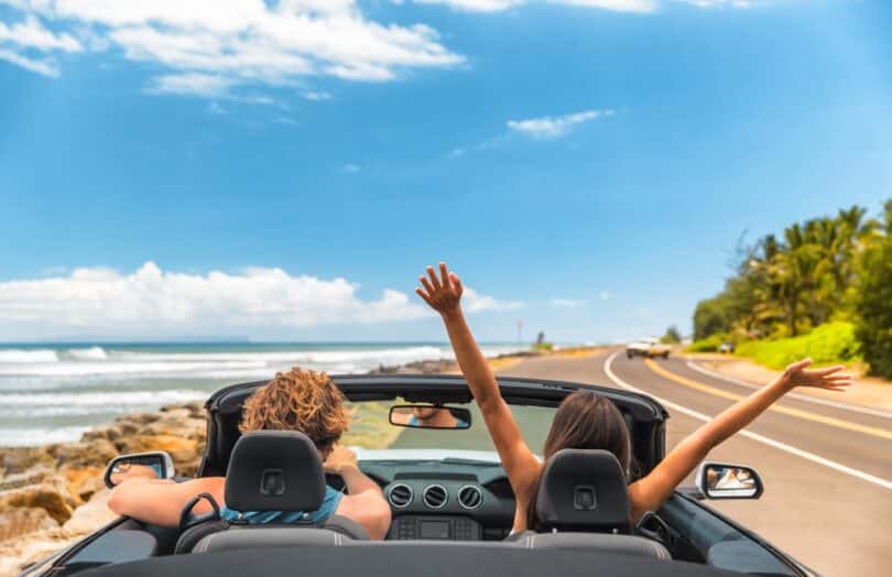 Casal de férias dentro de um conversível aberto na estrada. Na imagem há uma praia no fundo e a mulher está de braços abertos para cima