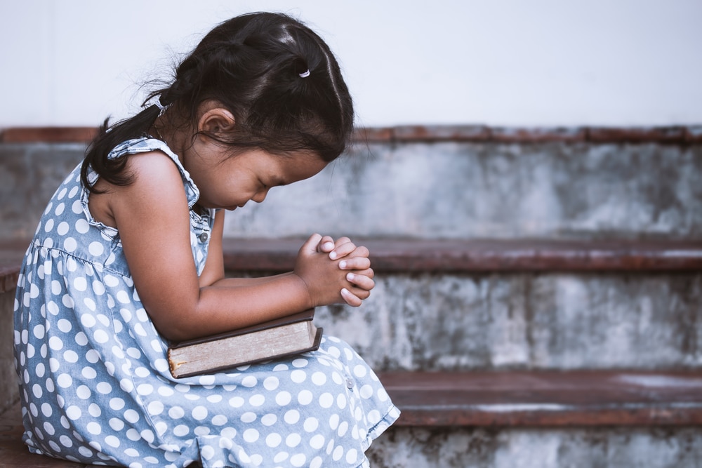 Menina criança de vestido azul com bolinhas brancas segurando uma Bíblia no colo e rezando com olhos fechados, cabeça baixa e mãos juntas