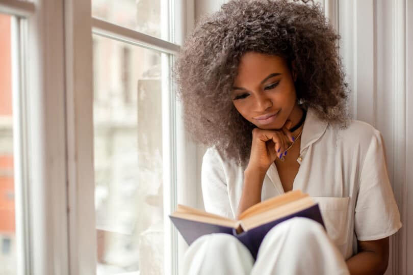 Mulher sentada na janela com mão esquerda no rosto e mão direita segurando livro