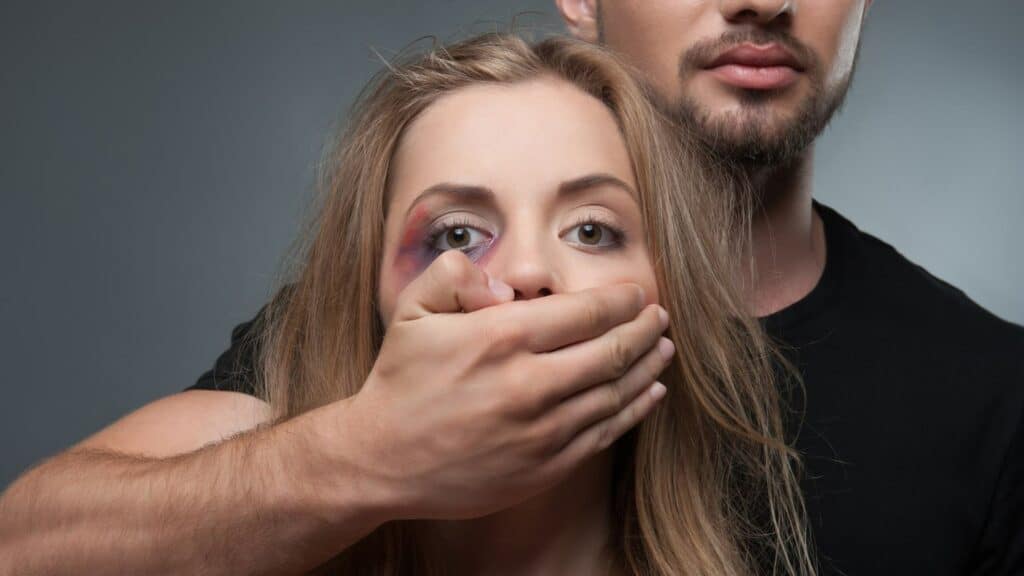 Um homem tapando a boca de uma mulher cujo lado direito da face apresenta uma roxidão intensa.