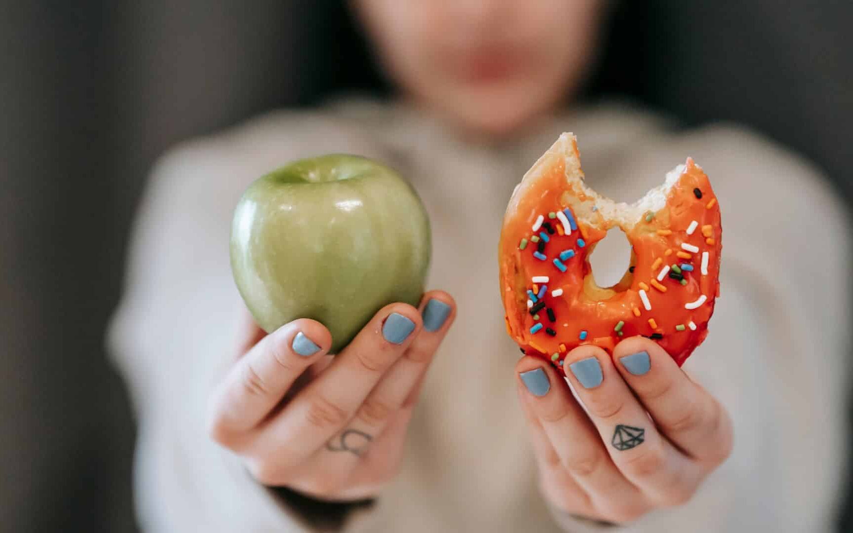 Em mãos diferentes, uma mulher segura uma rosquinha e uma maçã verde