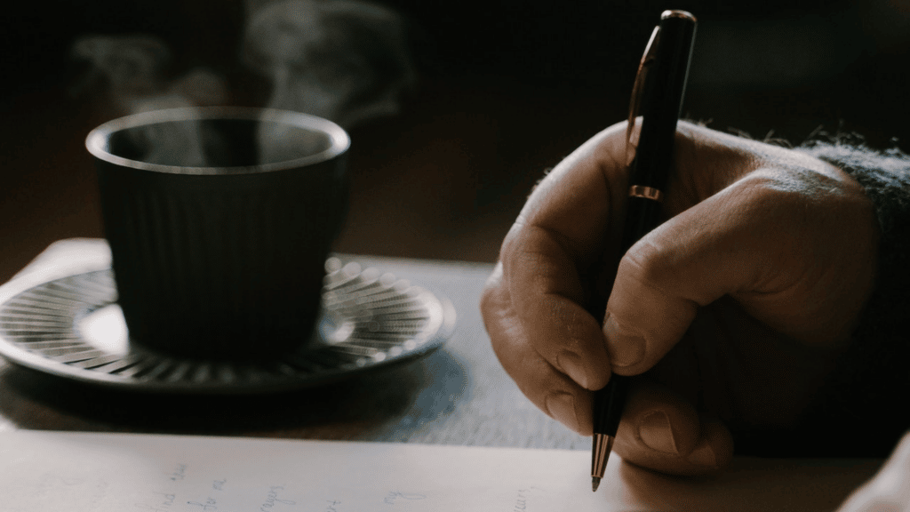 Foto de uma mão apoiada à mesa segurando uma caneta em frente a um papel e uma xícara de café do lado.