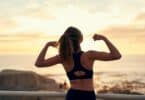 Uma mulher erguendo seus braços e exibindo seus bíceps à beira de uma praia.