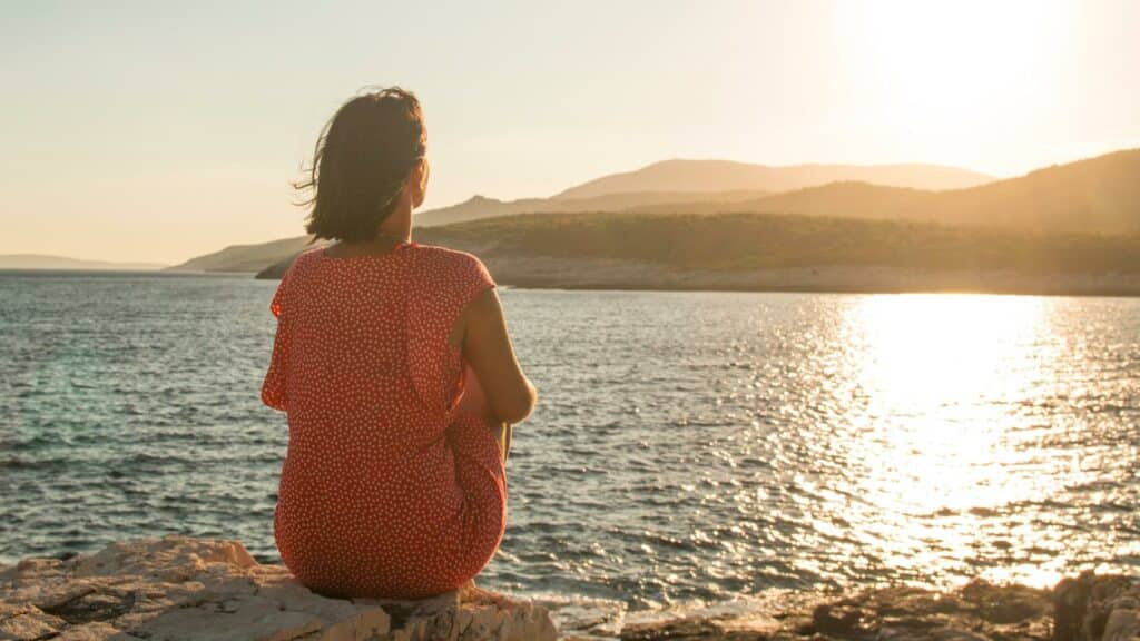 À beira de um extenso rio, uma mulher contemplando um sol cintilante à altura das montanhas.