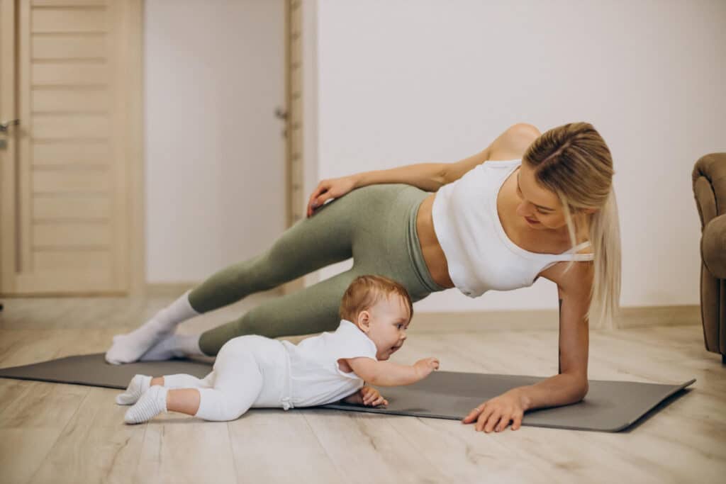 Uma mulher branca e loira fazendo uma posição de yoga enquanto seu filho engatinha ao lado dela.
