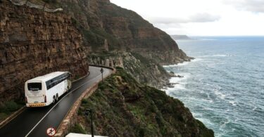 Foto de uma estrada um ônibus passando pela estrada e ao seu redor tem um penhasco e uma praia, como se fosse uma serra.