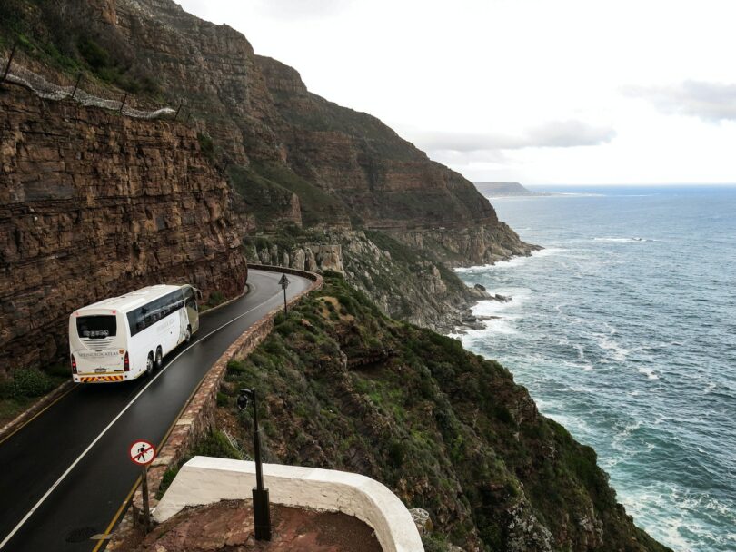 Foto de uma estrada um ônibus passando pela estrada e ao seu redor tem um penhasco e uma praia, como se fosse uma serra.