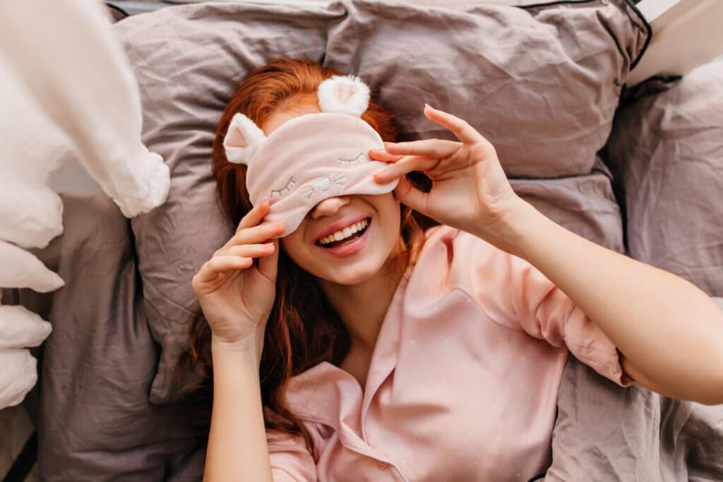 Mulher ruiva deitada na cama usando máscara para dormir, rindo.
